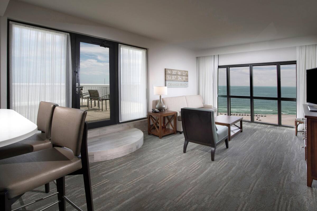 A hotel room with a large sliding glass door to the balcony, a flat-screen TV, a sofa, a dining table, and a small side table.