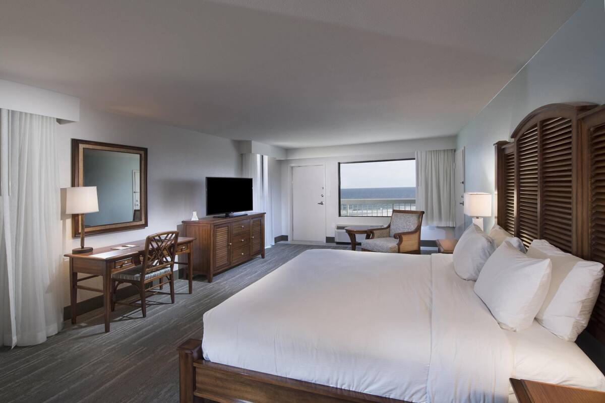 A hotel room with a large bed, a desk with a chair, a television, and a view of the beach.