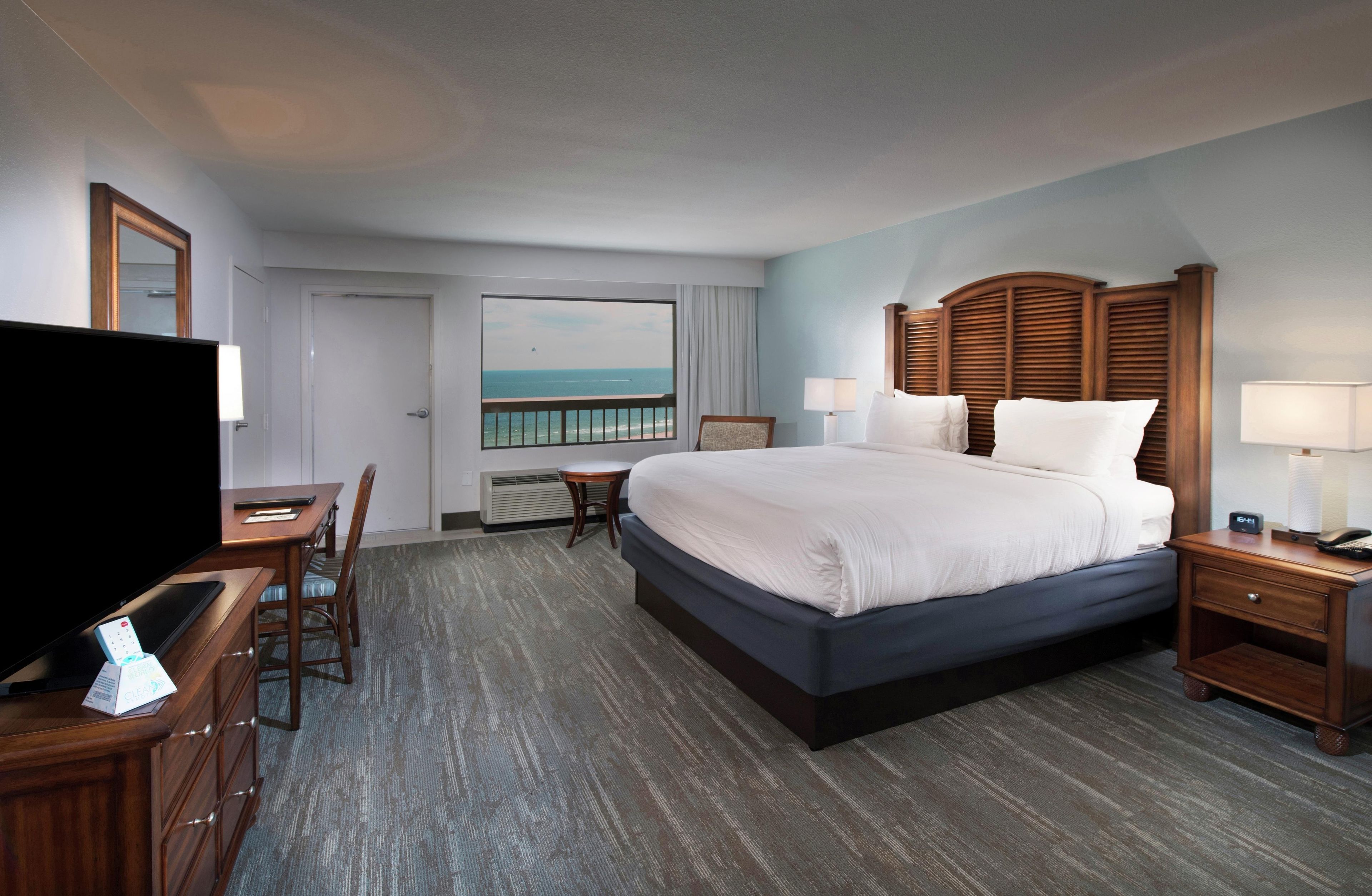 A hotel room with a large bed, a desk with a TV, a chair, a nightstand, and a view of the ocean.