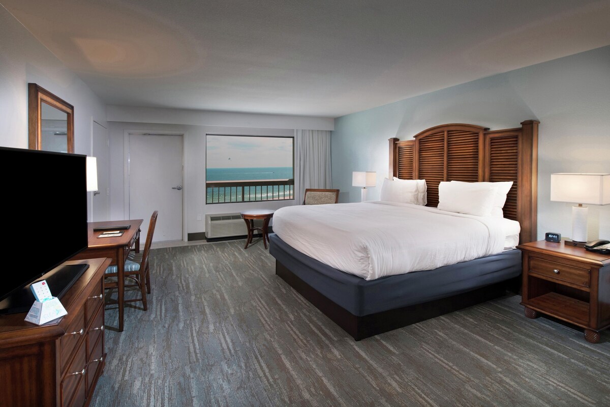 A hotel room with a large bed, a desk with a TV, a chair, a nightstand, and a view of the ocean.