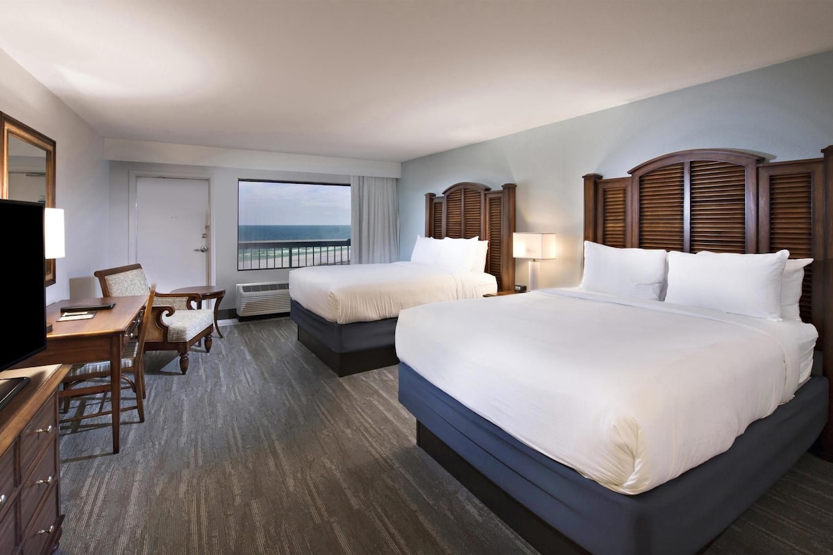 A hotel room with two beds, a desk, a chair, a television, and a window with a view of the ocean.