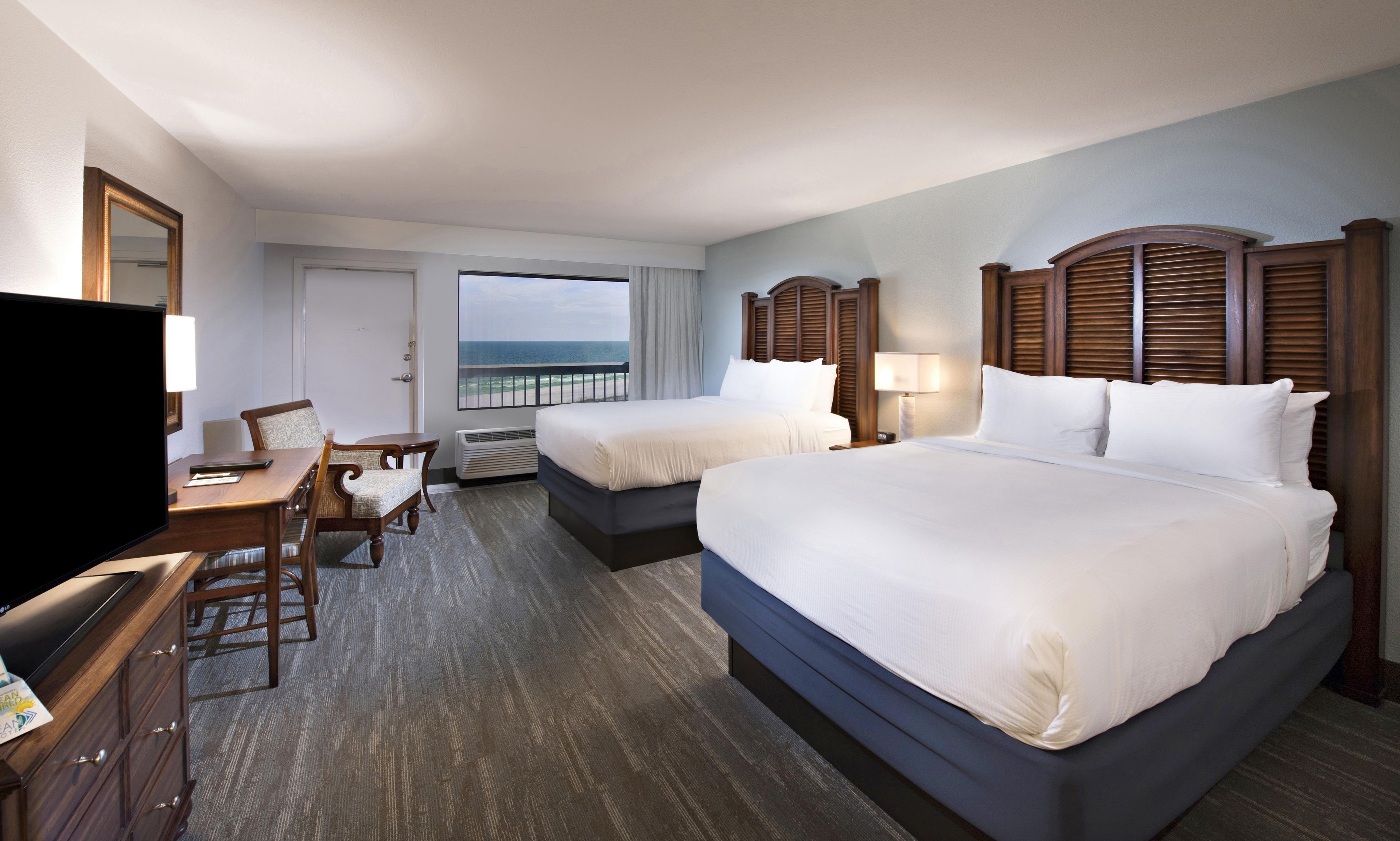 A hotel room with two beds, a desk, a chair, a TV, and a view of the ocean.