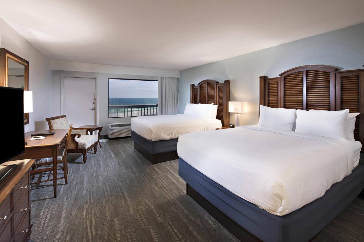 A hotel room with two beds, a desk, a chair, a TV, and a view of the ocean.