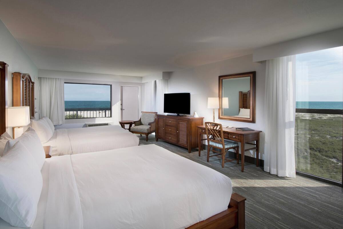 A hotel room with two beds, a TV, a desk with a chair, and a large window with a view of the ocean.