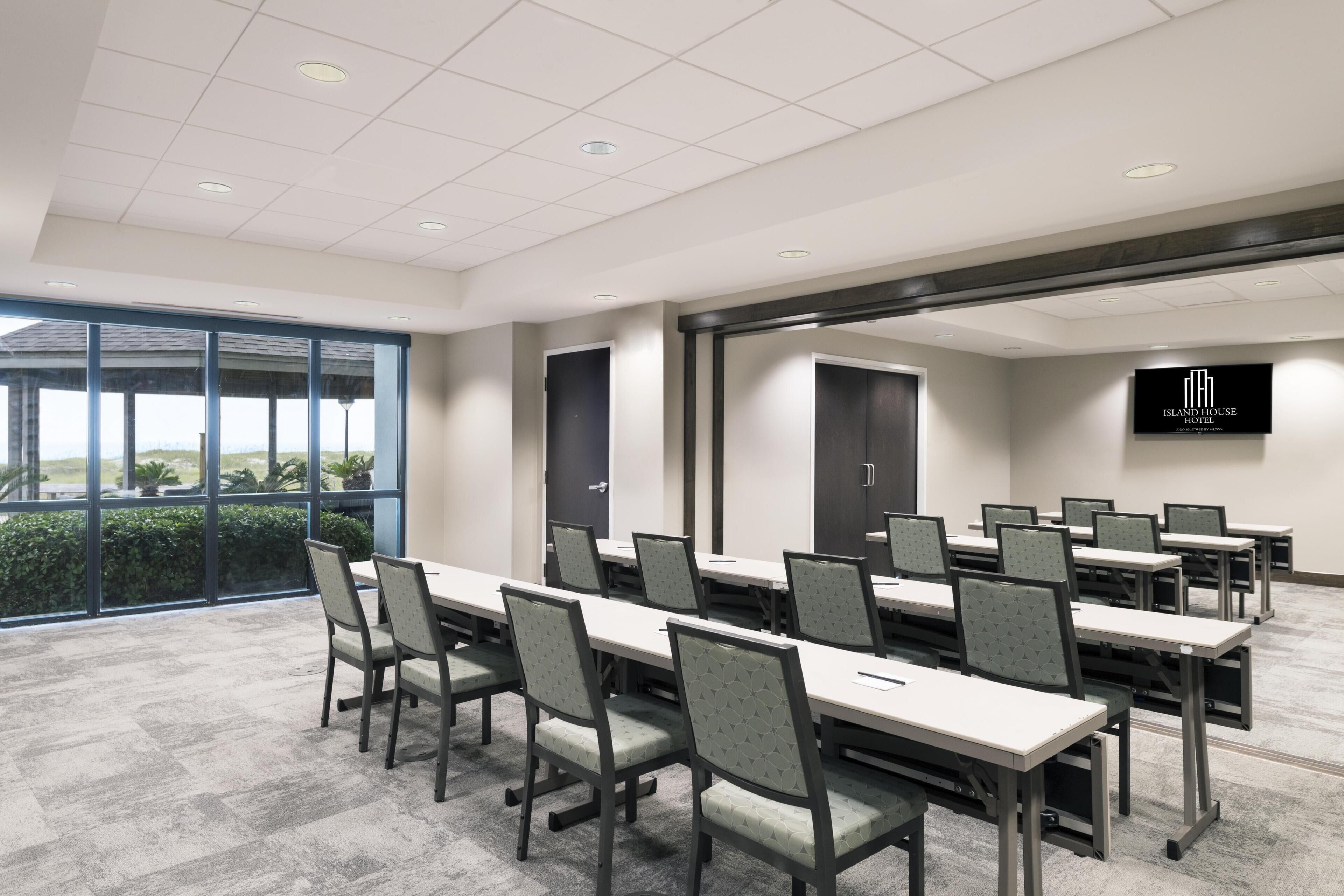 A conference room with long tables and chairs, a large window with a view, and a television screen displaying the hotel name.