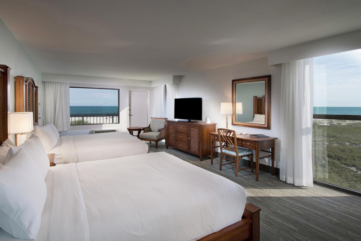 A hotel room with two beds, a TV, a desk with a chair, and a large window with a view of the ocean.