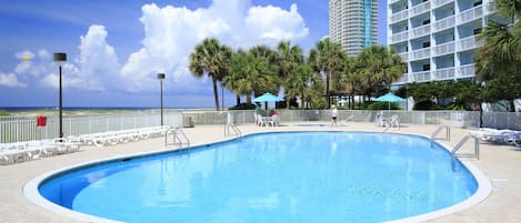 A swimming pool with a brick border, surrounded by palm trees and a tall building in the background.