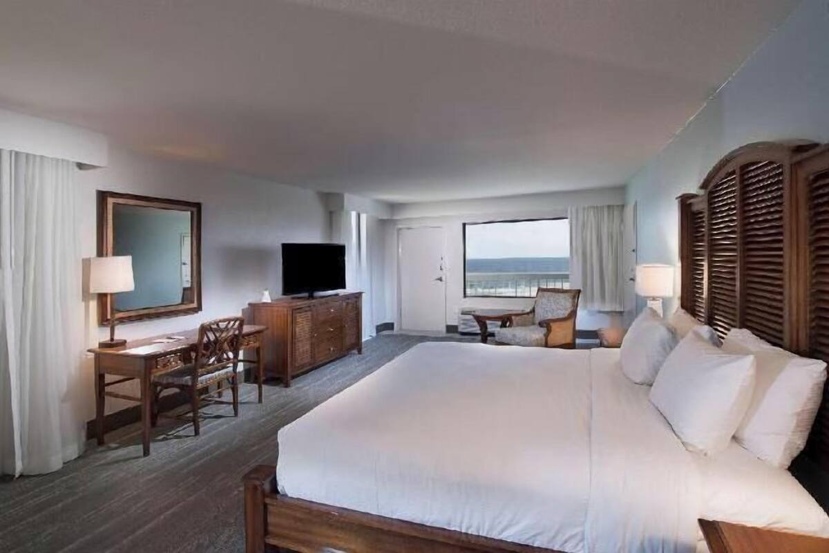A hotel room with a large bed, a desk, a chair, a television, and a view of the beach.