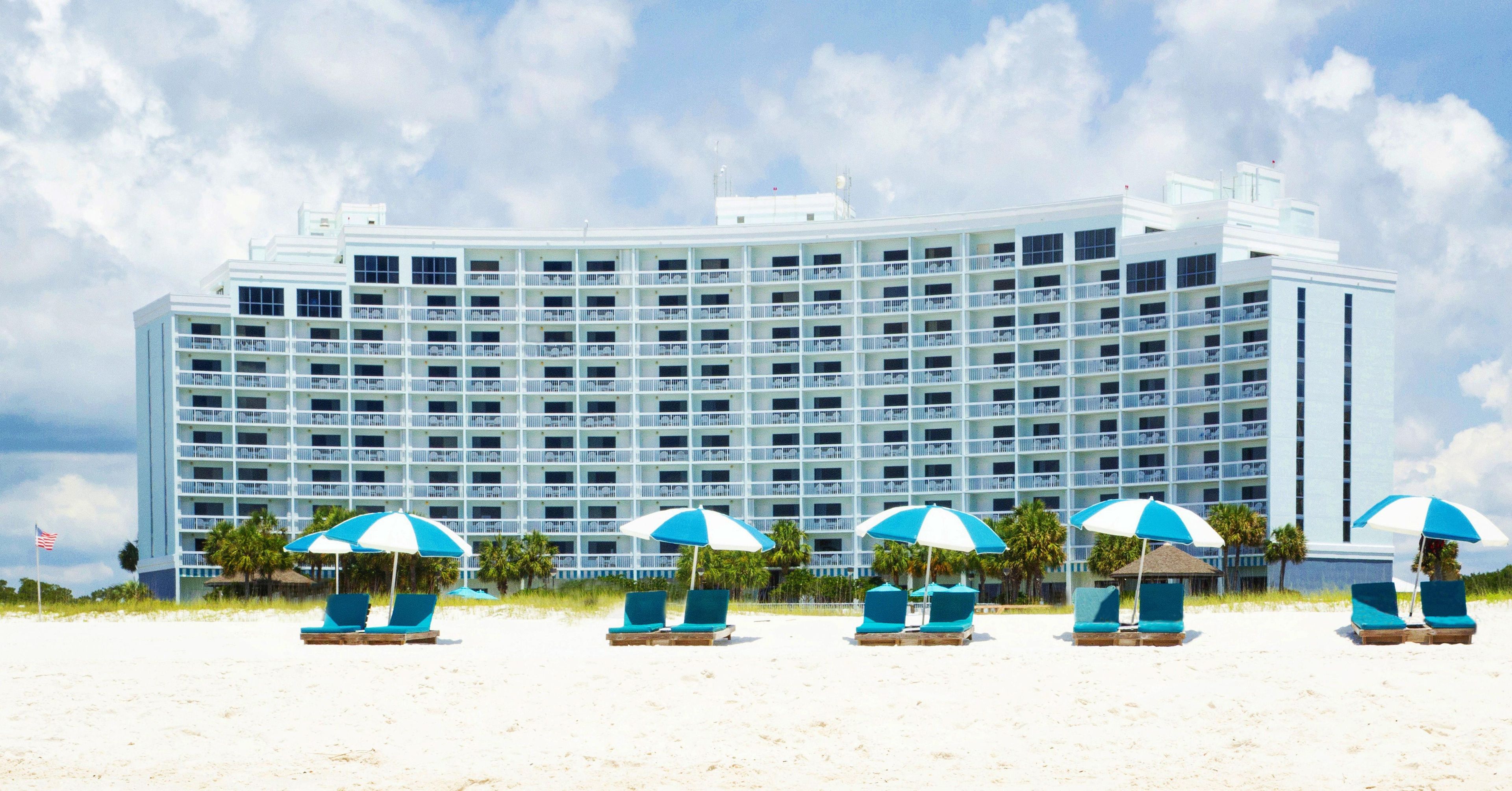 A large hotel with multiple balconies, a sandy beach in front, and several beach umbrellas.
