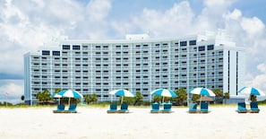 Un hotel grande con múltiples balcones, una playa de arena frente al establecimiento y varias sombrillas de playa.