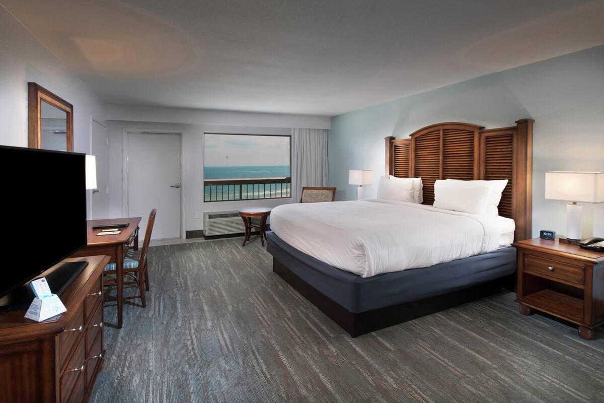 A hotel room with a large bed, a desk with a TV, a chair, a nightstand, and a view of the ocean.