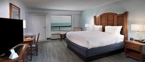 A hotel room with a large bed, a desk with a TV, a chair, a nightstand, and a view of the ocean.