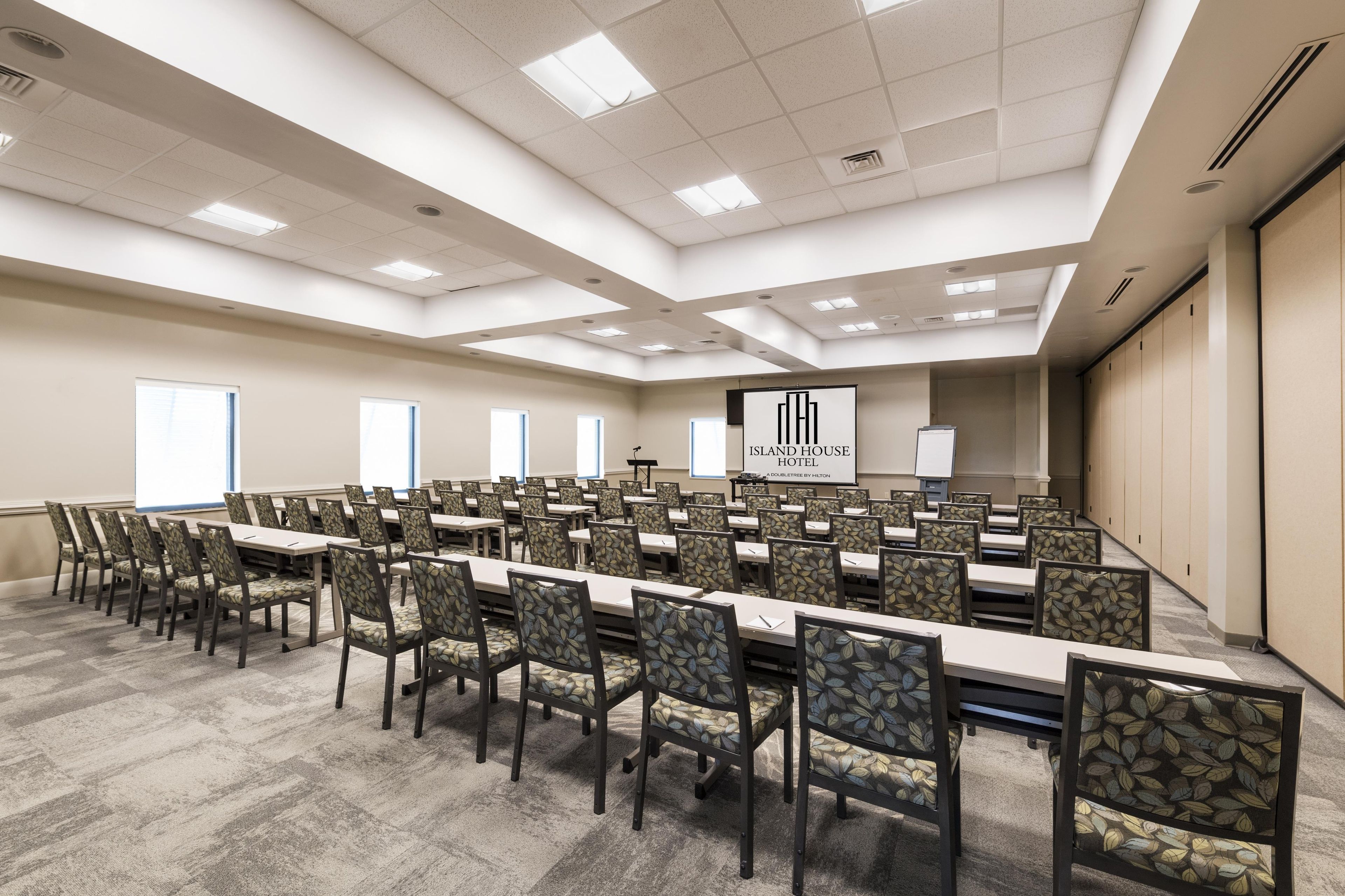 Una sala de conferencias con varias mesas y sillas, una pantalla grande mostrando el nombre del hotel, e iluminación empotrada en el techo.