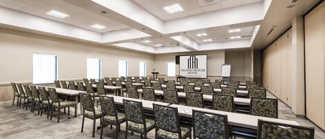 A conference room with multiple tables and chairs, a large screen displaying the hotel name, and a ceiling with recessed lighting.