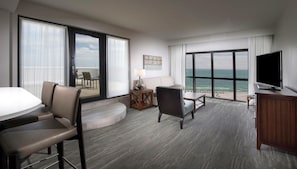 A hotel room with a large sliding glass door to the balcony, a flat-screen TV, a sofa, a dining table, and a small side table.
