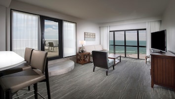 A hotel room with a large sliding glass door to the balcony, a flat-screen TV, a sofa, a dining table, and a small side table.