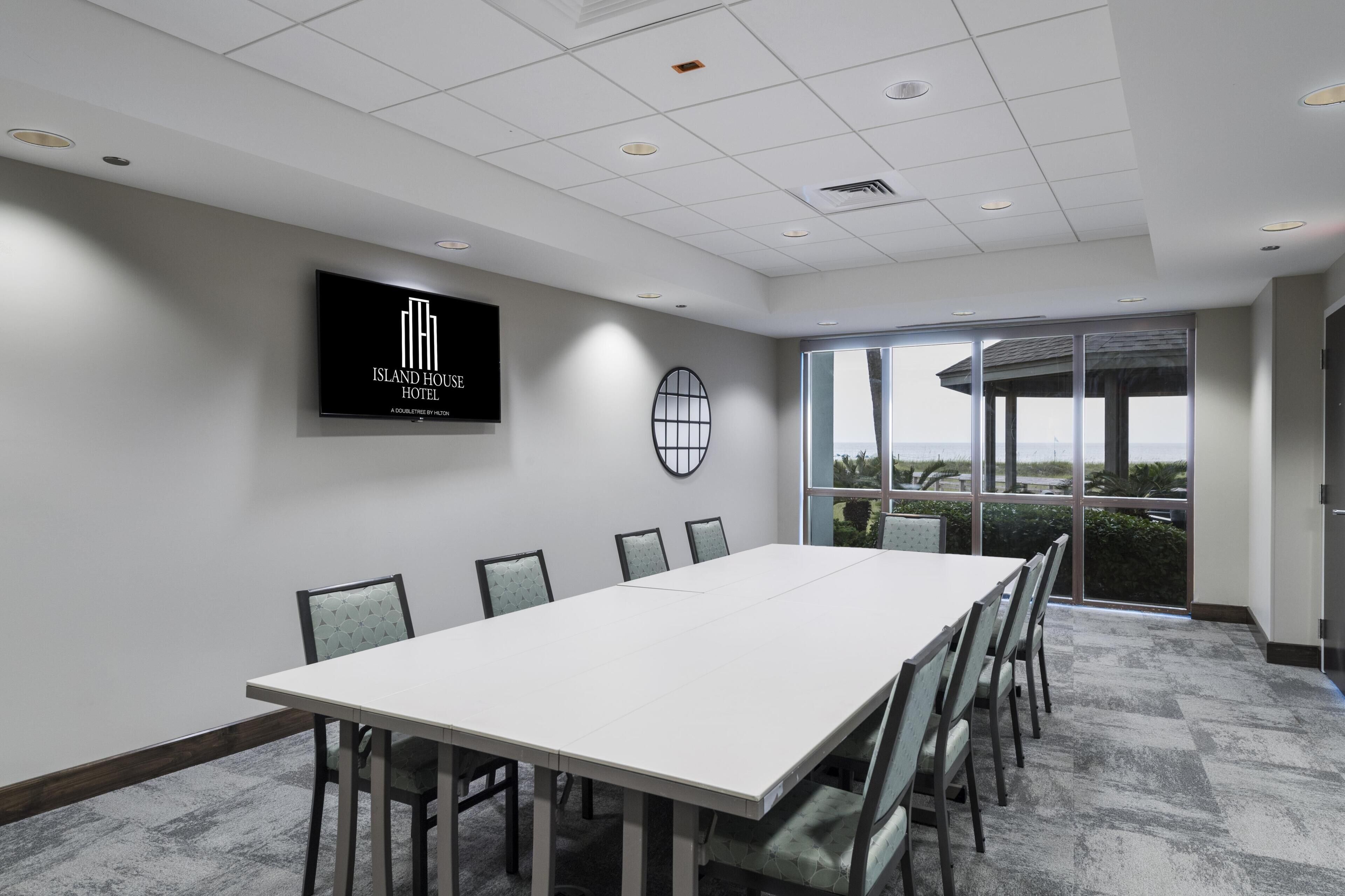 A conference room with a long table, chairs, a TV screen with the hotel logo, a large window with a view, and a ceiling with recessed lighting.
