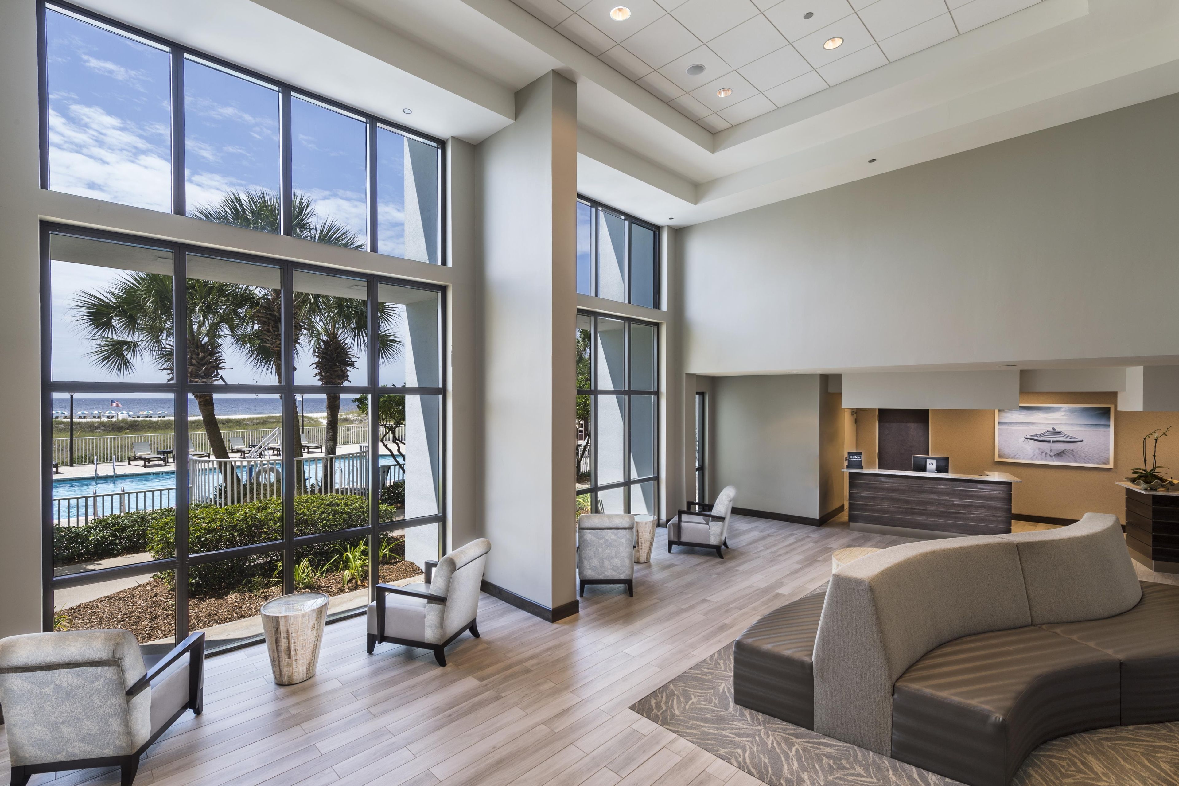 A spacious lobby with large windows, a reception desk, and a seating area with a view of the pool and palm trees.