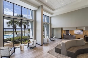 Un vestíbulo amplio con ventanas grandes, una recepción y un área de descanso con vista a la piscina y palmeras.