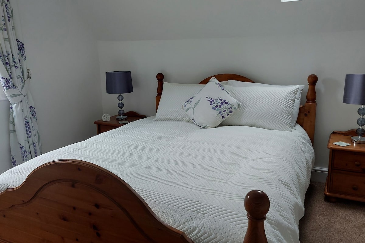 A neatly made bed with a wooden headboard, flanked by two bedside tables with lamps, in a simple room with plain walls and a window with curtains.