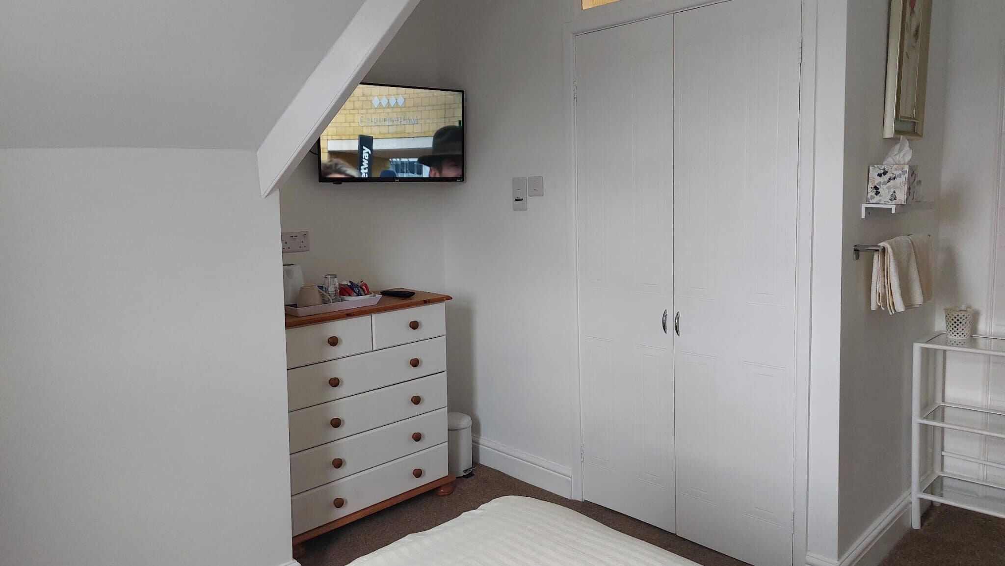 A bedroom with a white wardrobe, a wooden dresser, a TV mounted on the wall, and a white bedside table.