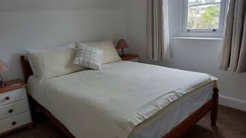 Una cama bien hecha con edredón blanco y dos almohadas, una mesita de noche de madera con una lámpara y una ventana con cortinas.