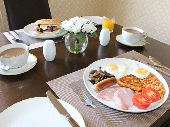 A breakfast setting with a plate of eggs, sausages, tomatoes, and beans, a cup of coffee, and a glass of orange juice on a wooden table.