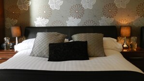 Een opgemaakt bed met een zwart hoofdeinde, twee witte kussens en twee kussens met een patroon. Aan beide zijden van het bed staan nachtlampjes.