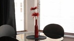 Een ronde glazen tafel met een rode vaas met rode bloemen, twee zwarte stoelen, en een raam met jaloezieën op de achtergrond.