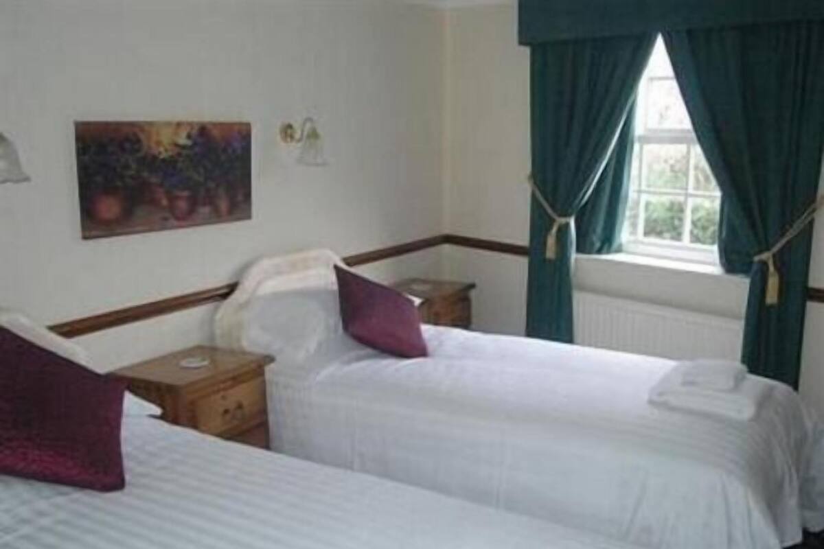 A hotel room with two single beds, a nightstand, a lamp, a painting on the wall, and a window with curtains.