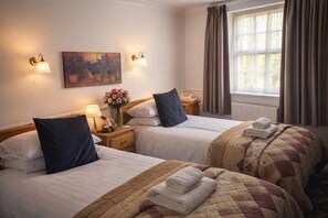 Ett hotellrum med två sängar, en tavla, vägglampor, ett fönster med gardiner och ett nattduksbord med en lampa och blommor.