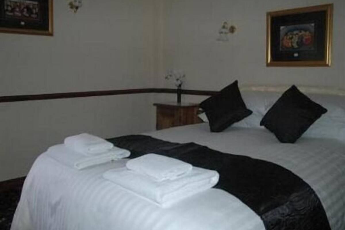 A neatly made bed with white linens and black pillows, a small wooden nightstand with a vase of flowers, and framed artwork on the walls.