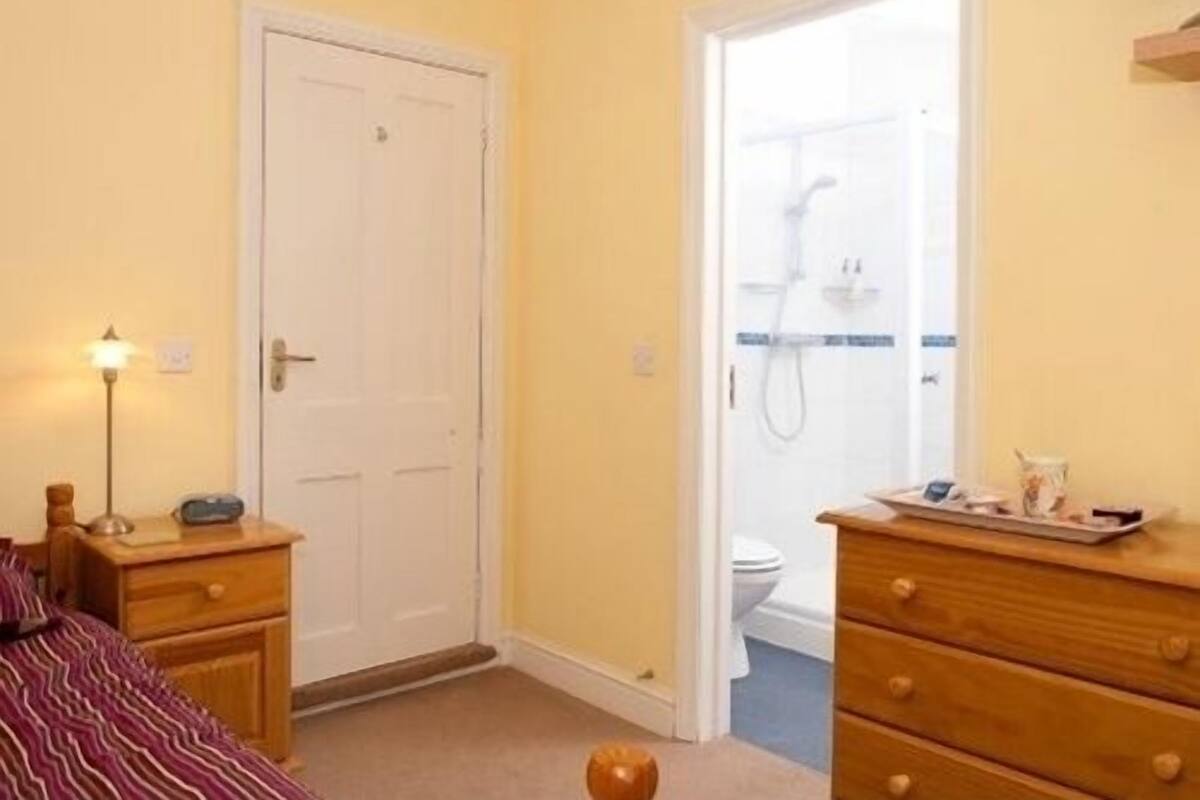 A bedroom with a bed, a bedside table, a dresser, and a door leading to a bathroom with a shower.