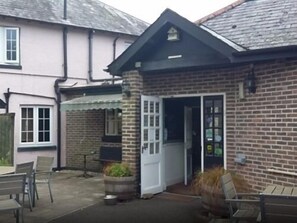 A brick building with a white door and windows, a small outdoor seating area with chairs, and a potted plant. - The Brewers Arms (Dorchester)