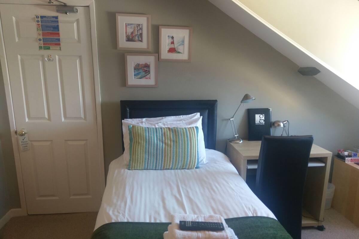 A neatly arranged bedroom with a bed, a desk, and a chair. There are framed pictures on the wall and a door with a sign.