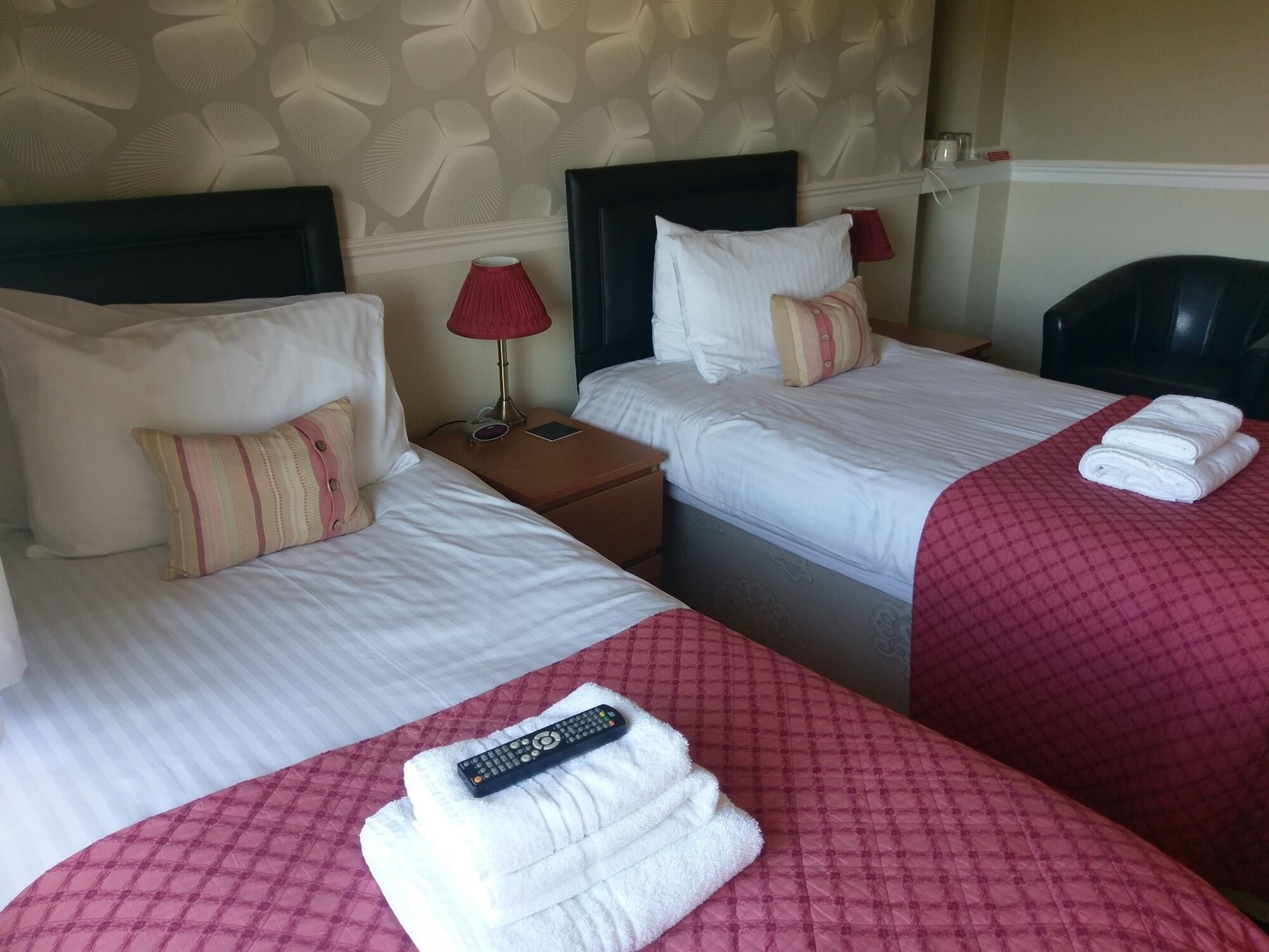 A hotel room with two beds, each with white linens and red blankets. There are two bedside tables, one with a lamp and the other with a remote control.