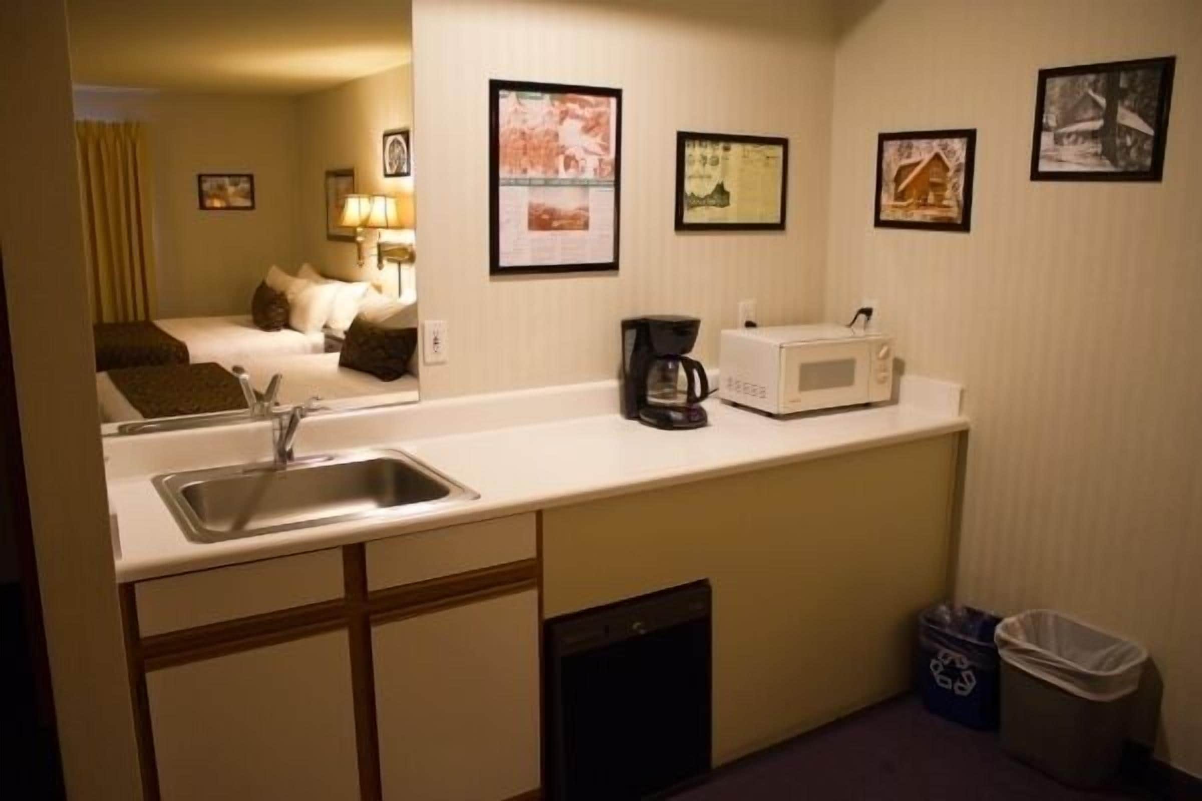 A compact hotel room with a kitchenette, a bed, a mirror, and framed pictures on the wall.