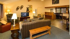 A cozy living room with a sofa, a television, and a wooden bench. There is a dining area with a table and chairs, and a kitchen in the background. - Holiday Haus Motel (Mammoth Lakes)