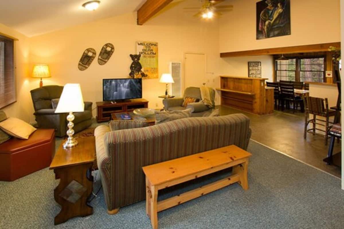 A cozy living room with a sofa, a television, and a wooden bench. There is a dining area with a table and chairs, and a kitchen in the background.