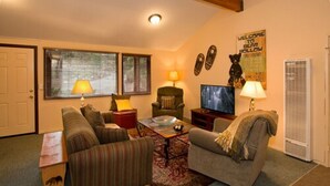A cozy living room with a sofa, armchairs, a coffee table, and a television. There are two lamps, a rug, and a door. - Holiday Haus Motel (Mammoth Lakes)
