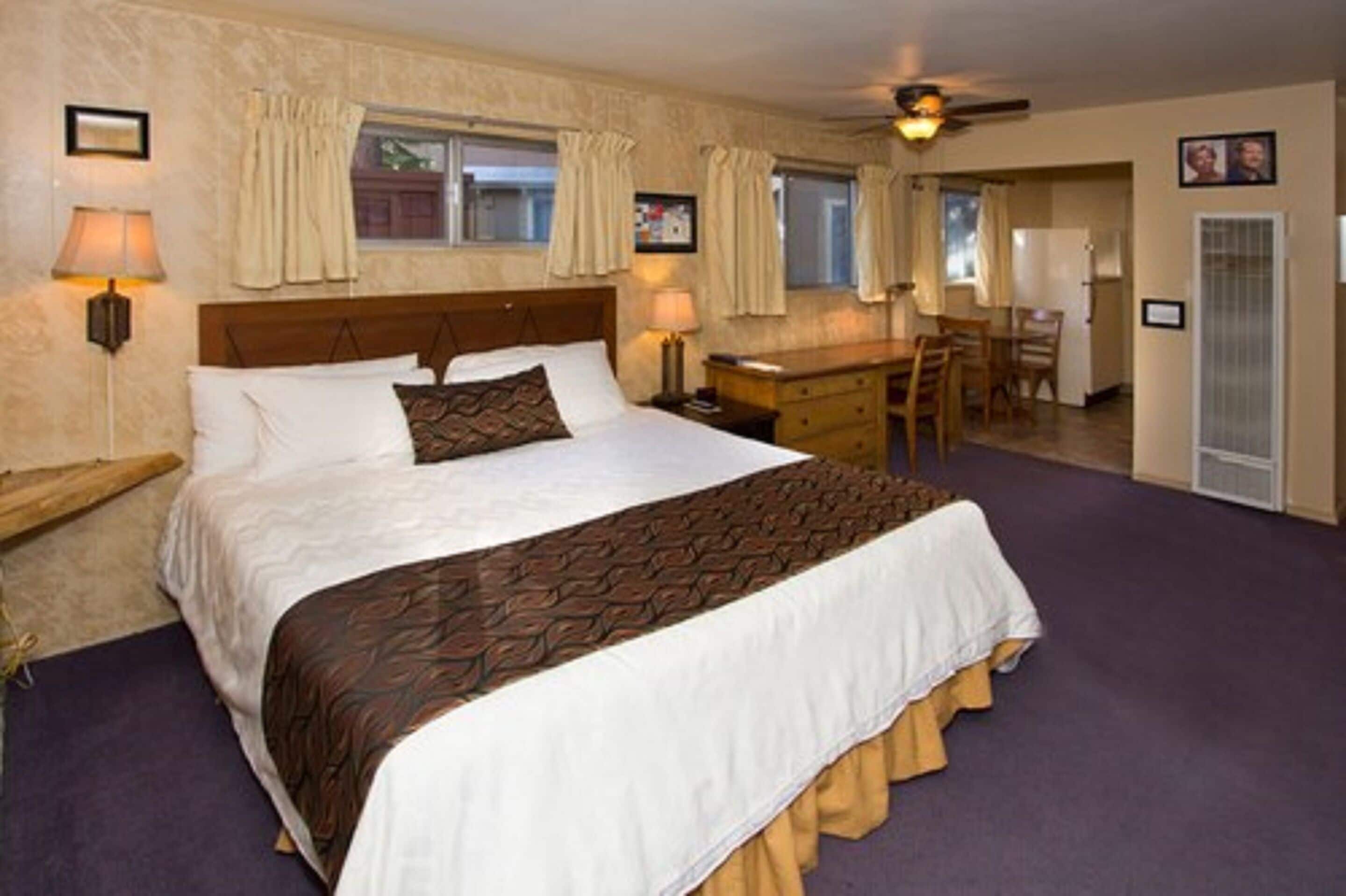 A hotel room with a large bed, a desk, and a chair. There are two windows with curtains, a ceiling fan, and a door with a picture.