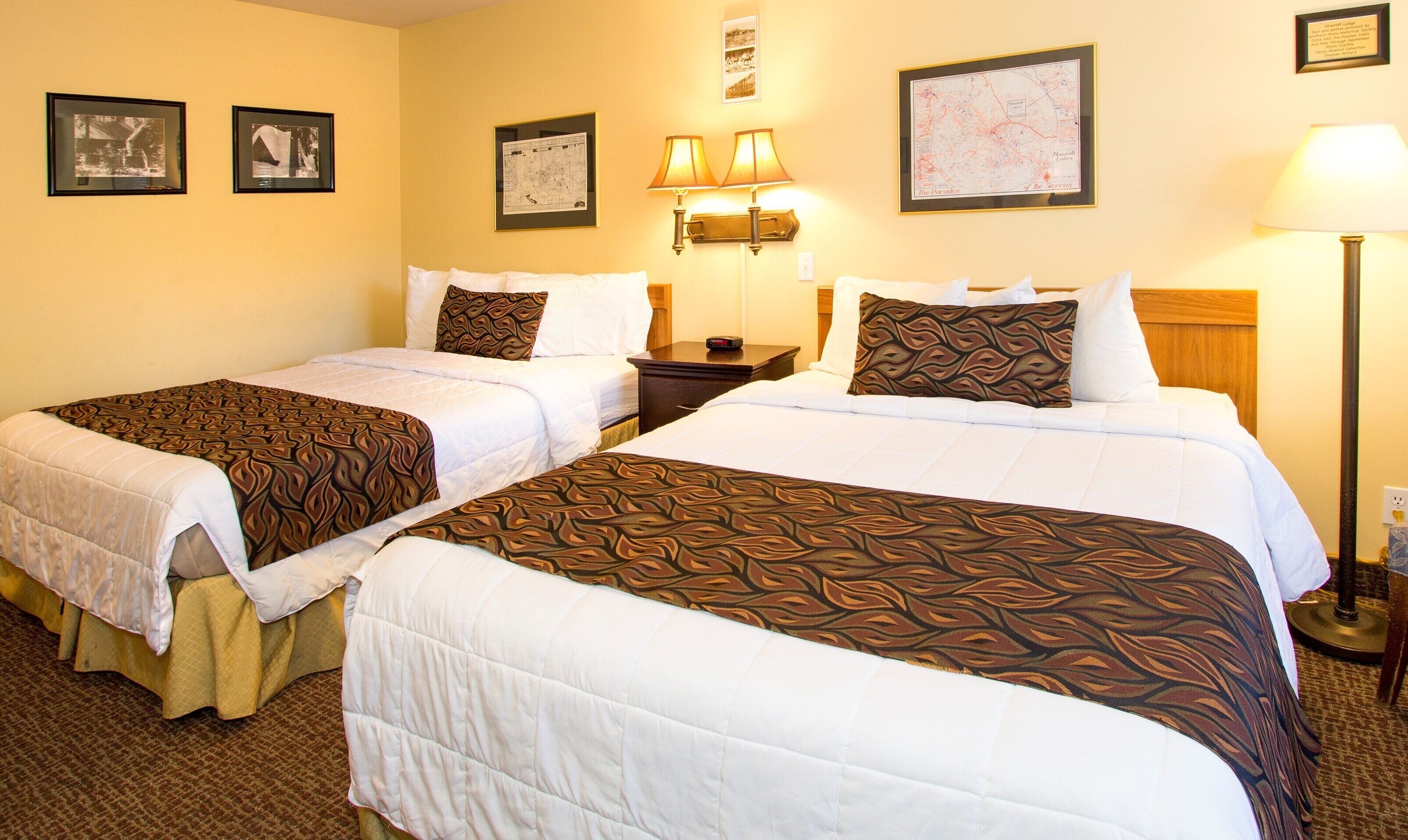 A hotel room with two beds, each with a patterned bedspread, a nightstand with a lamp, and framed pictures on the walls.