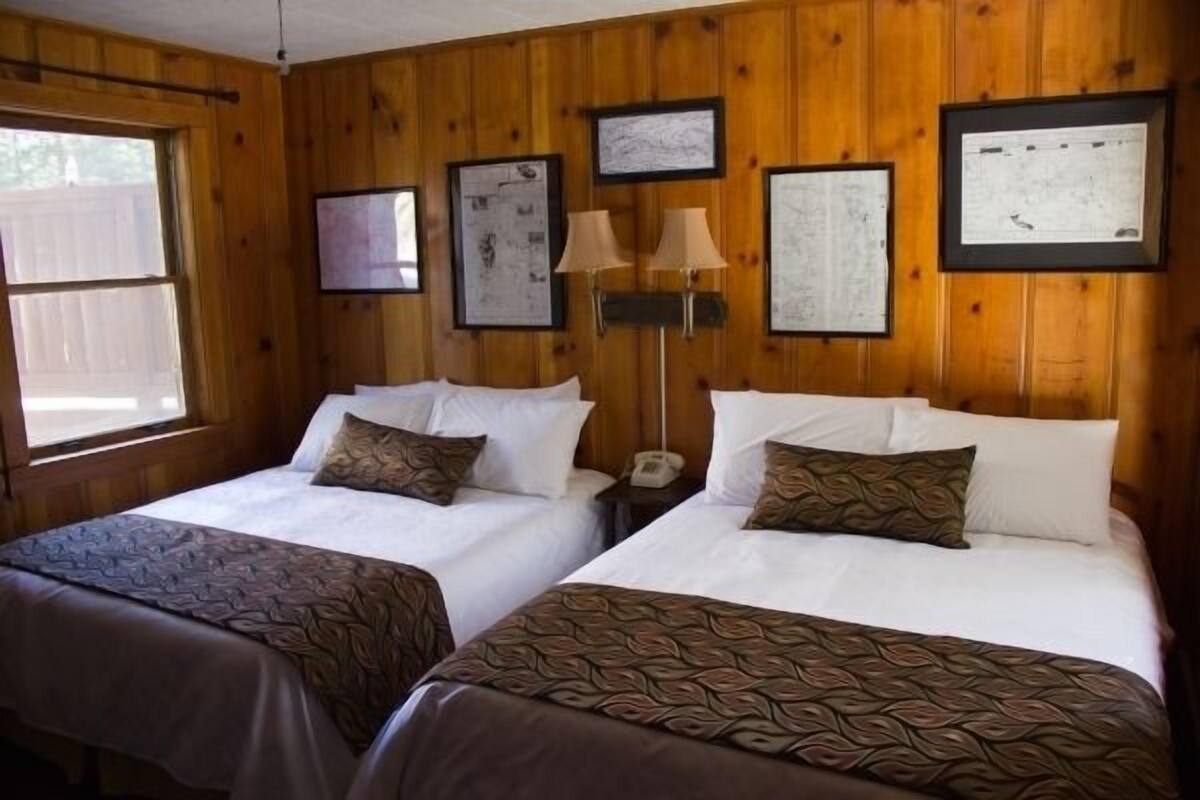 A room with two beds, wooden walls, framed pictures, and a window.