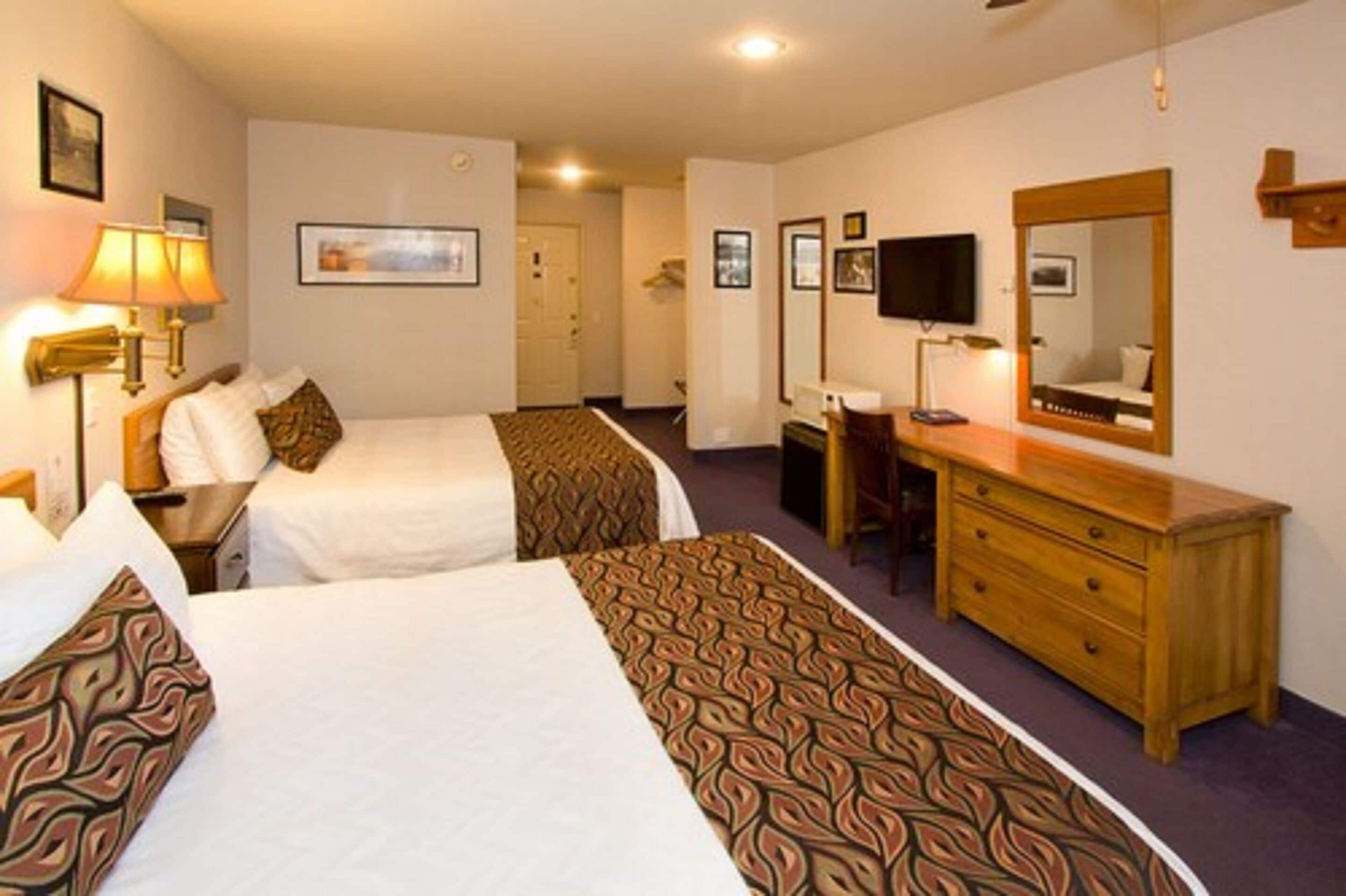A hotel room with two beds, a desk, a dresser, a television, and a mirror.
