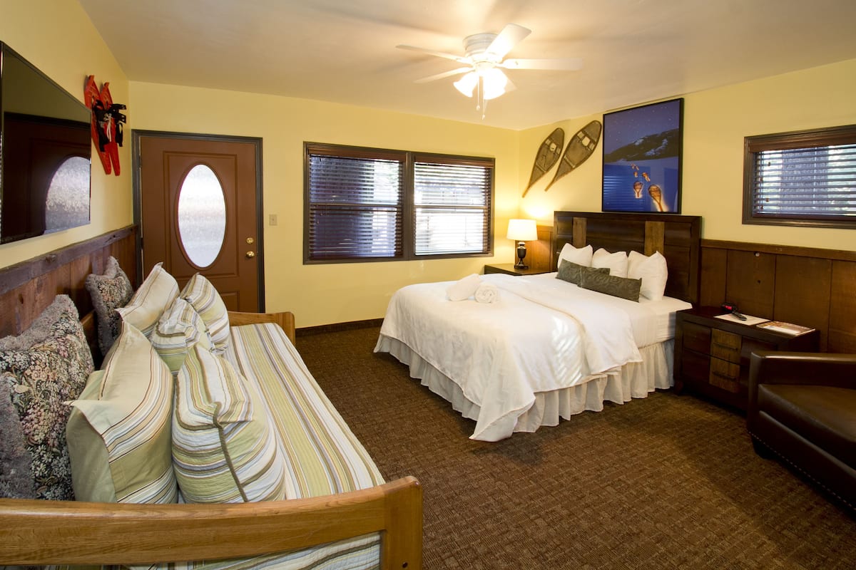 A hotel room with a bed, a sofa, a wooden headboard, a nightstand, a lamp, and a ceiling fan.