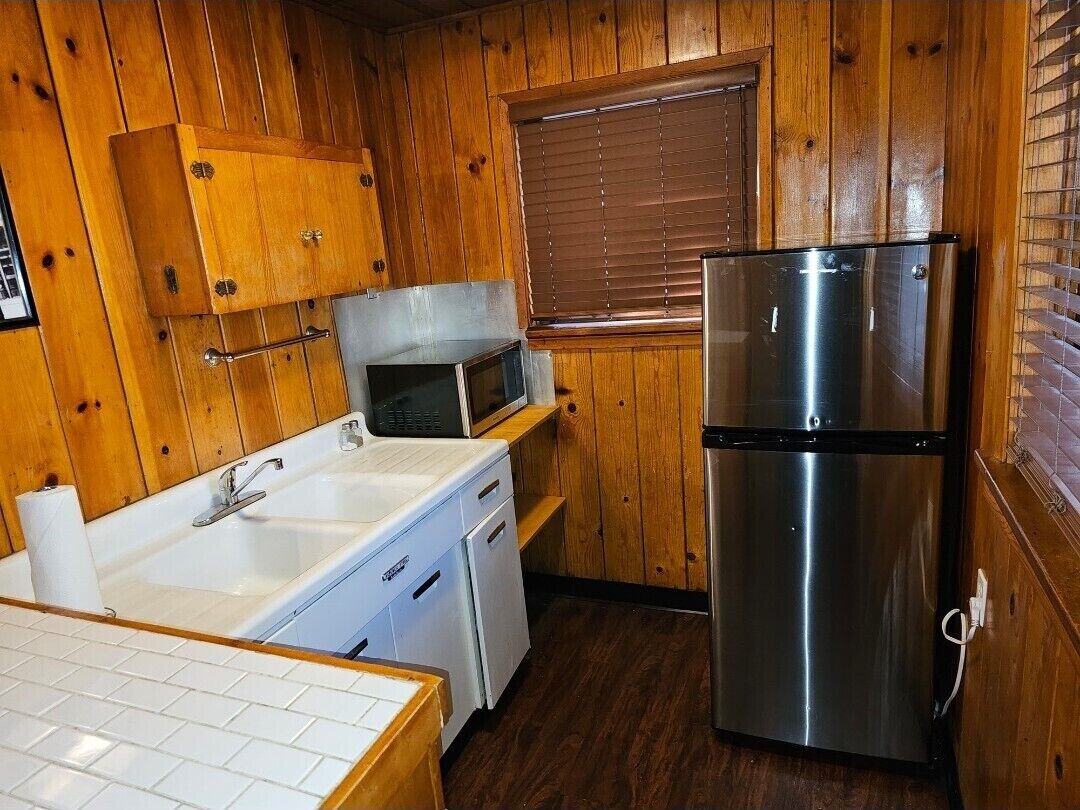 Cocina compacta con paredes de madera, un refrigerador de acero inoxidable, un microondas y un lavamanos con encimera blanca.