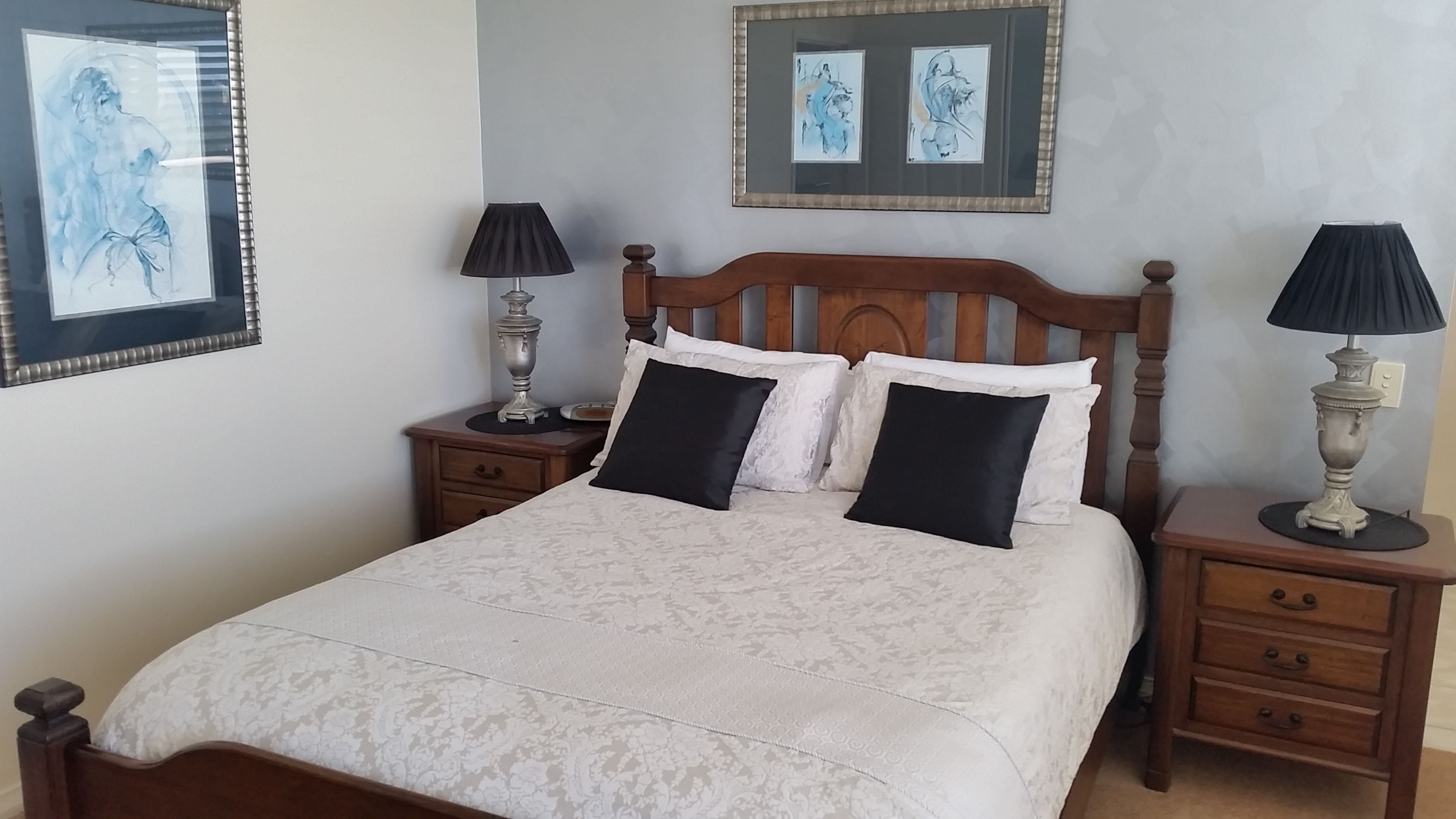 A neatly made bed with a white duvet and black pillows, flanked by two bedside tables with lamps, in a room with artwork on the walls.