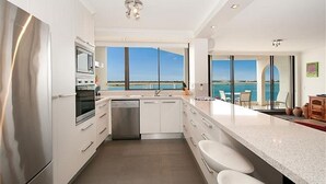 A modern kitchen with a view of the sea, stainless steel appliances, and a large island with seating. - Beaconlea Tower Apartments (Labrador)