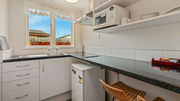 A compact kitchen with white cabinets, a microwave, and a small refrigerator. There is a window with a view of a house and a clear sky. - Arcadia Motel (Christchurch)
