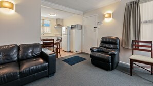A living room with a black leather sofa, a black leather chair, a wooden chair, a blue rug, and a kitchen in the background. - Arcadia Motel (Christchurch)